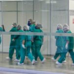 Profissionais de saúde caminhando em um corredor do hospital com roupas hospitalares (traje na cor azul, touca de cabelo e máscara). Eles são os principais "alvos" dos transtornos mentais.