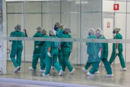 Profissionais de saúde caminhando em um corredor do hospital com roupas hospitalares (traje na cor azul, touca de cabelo e máscara). Eles são os principais "alvos" dos transtornos mentais.