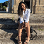 Mulher parda, jovem, com cabelos escuros cacheados, vestindo uma camisa branca e shorts escuros, sentada em degraus de pedra com um edifício antigo ao fundo.