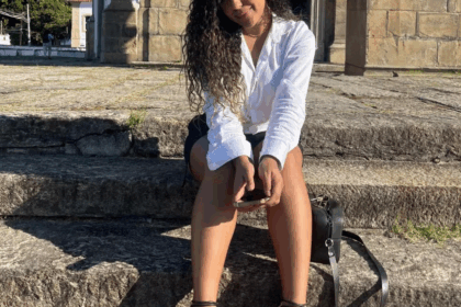 Mulher parda, jovem, com cabelos escuros cacheados, vestindo uma camisa branca e shorts escuros, sentada em degraus de pedra com um edifício antigo ao fundo.