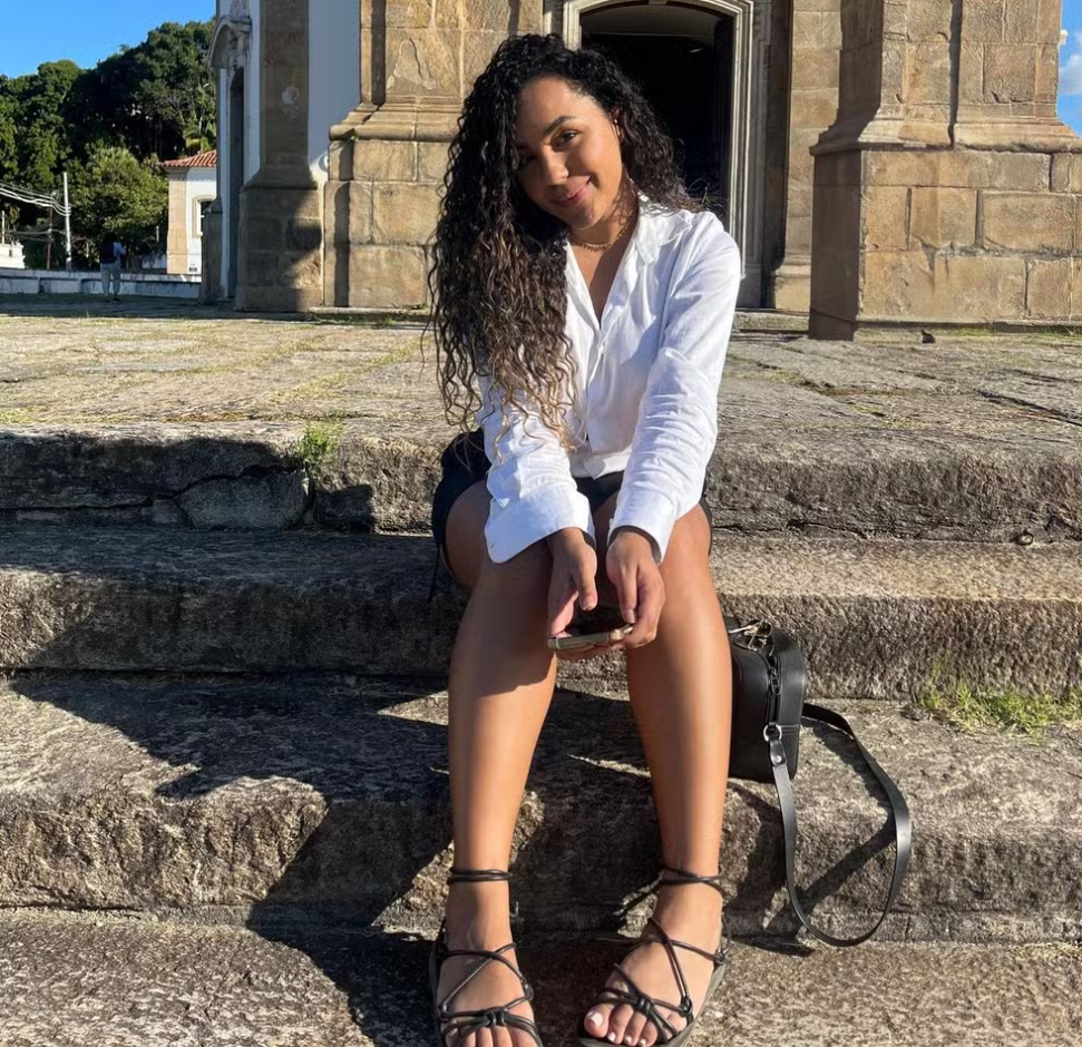 Mulher parda, jovem, com cabelos escuros cacheados, vestindo uma camisa branca e shorts escuros, sentada em degraus de pedra com um edifício antigo ao fundo.