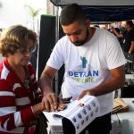 Ilustrar o atendimento do programa Detran Presente em ação, mostrando usuários sendo atendidos no Ciep de São João de Meriti, para reforçar a proximidade e acessibilidade dos serviços do Detran RJ oferecidos gratuitamente ou com facilidade de pagamento, destacando a iniciativa de levar documentação e regularização veicular para a população local.