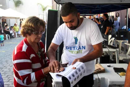 Ilustrar o atendimento do programa Detran Presente em ação, mostrando usuários sendo atendidos no Ciep de São João de Meriti, para reforçar a proximidade e acessibilidade dos serviços do Detran RJ oferecidos gratuitamente ou com facilidade de pagamento, destacando a iniciativa de levar documentação e regularização veicular para a população local.