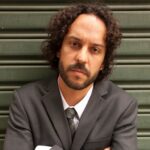 Imagem do rapper Gabriel O Pensador, de braços cruzados, vestindo um terno cinza e em frente a uma parede metálica de tom verde. O artista está com o cabelo médio (até o queixo), cavanhaque e uma feição séria.