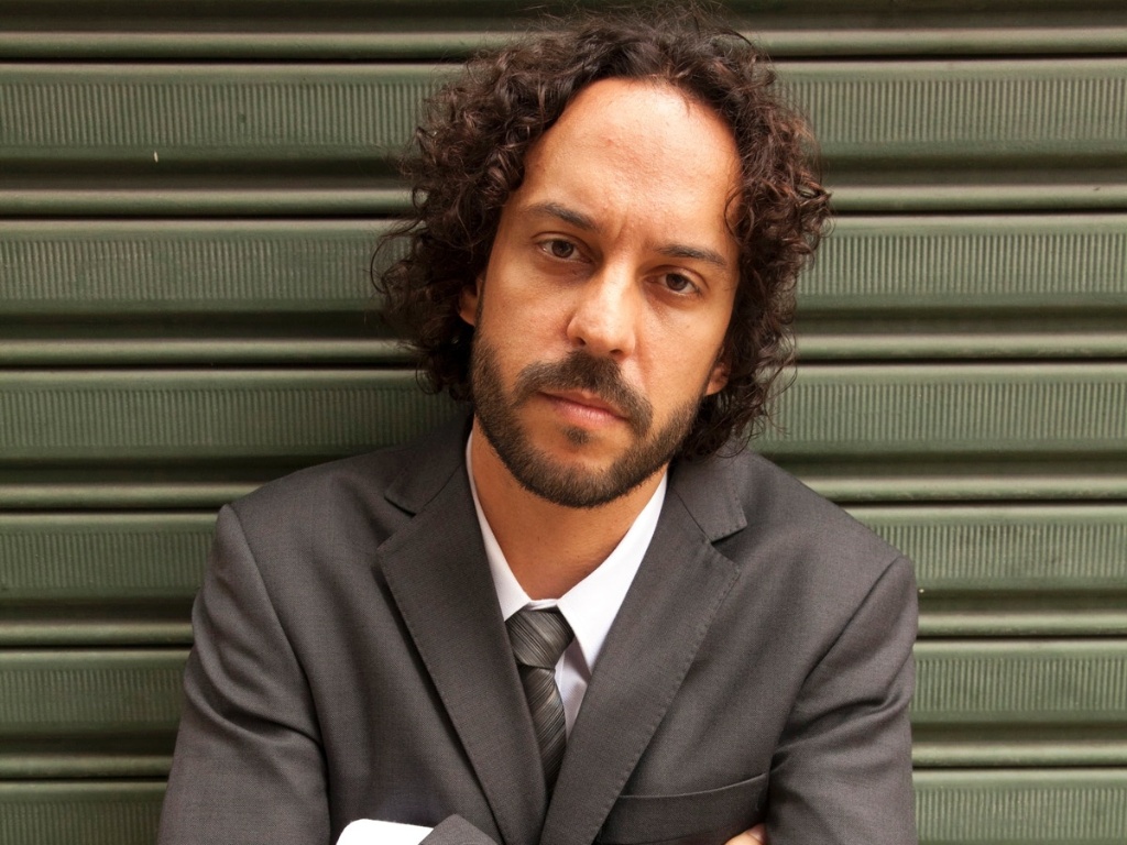 Imagem do rapper Gabriel O Pensador, de braços cruzados, vestindo um terno cinza e em frente a uma parede metálica de tom verde. O artista está com o cabelo médio (até o queixo), cavanhaque e uma feição séria.