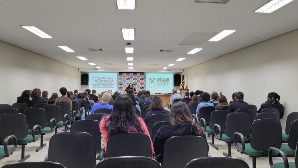 Auditório da UniSantos durante o Culturalmente Santista 2024, com público assistindo a uma palestra. A imagem mostra cadeiras ocupadas e o telão exibindo o logotipo do evento “Culturalmente Santista – Fórum Cultural e Criativo de Santos”.
