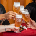 Quatro pessoas brindam com taças de cerveja artesanal, marcadas com o logotipo da Academia da Cerveja. As bebidas apresentam uma coloração dourada e espuma cremosa, em um ambiente acolhedor, com mesa coberta por uma toalha vermelha e detalhes de decoração simples, como flores secas e utensílios de mesa. A cena transmite celebração, amizade e valorização dos sabores brasileiros — especialmente aqueles inspirados nos ingredientes e na cultura do Nordeste.