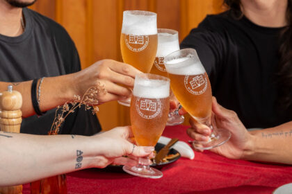 Quatro pessoas brindam com taças de cerveja artesanal, marcadas com o logotipo da Academia da Cerveja. As bebidas apresentam uma coloração dourada e espuma cremosa, em um ambiente acolhedor, com mesa coberta por uma toalha vermelha e detalhes de decoração simples, como flores secas e utensílios de mesa. A cena transmite celebração, amizade e valorização dos sabores brasileiros — especialmente aqueles inspirados nos ingredientes e na cultura do Nordeste.