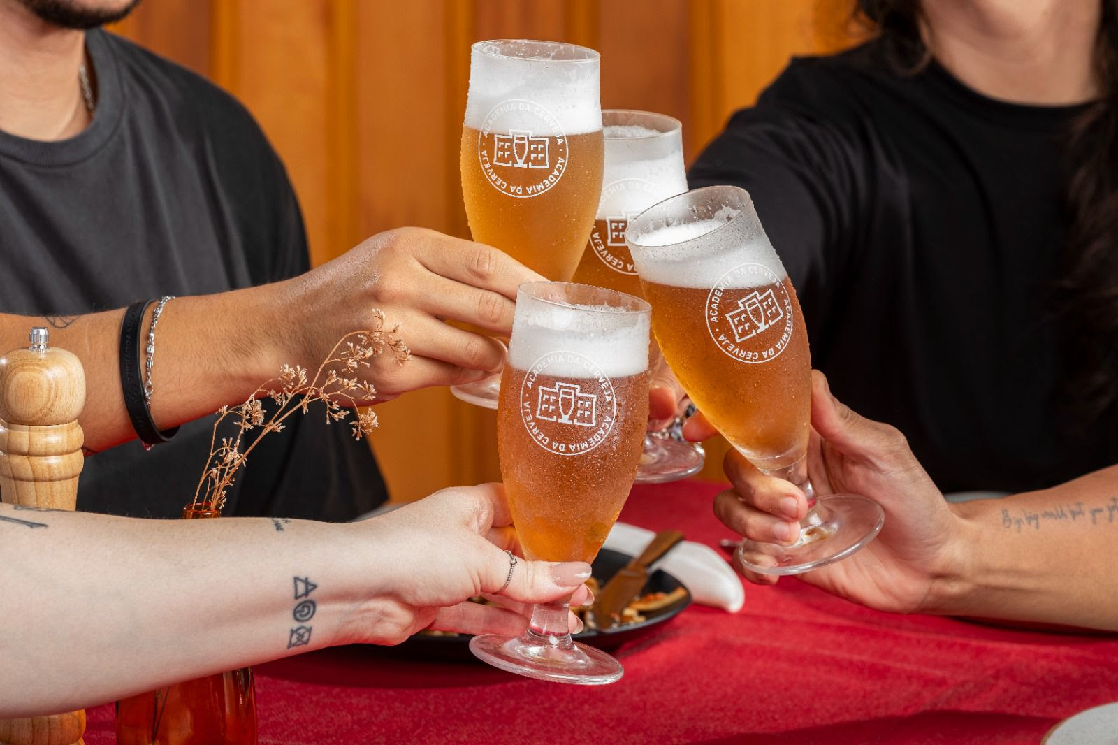 Quatro pessoas brindam com taças de cerveja artesanal, marcadas com o logotipo da Academia da Cerveja. As bebidas apresentam uma coloração dourada e espuma cremosa, em um ambiente acolhedor, com mesa coberta por uma toalha vermelha e detalhes de decoração simples, como flores secas e utensílios de mesa. A cena transmite celebração, amizade e valorização dos sabores brasileiros — especialmente aqueles inspirados nos ingredientes e na cultura do Nordeste.