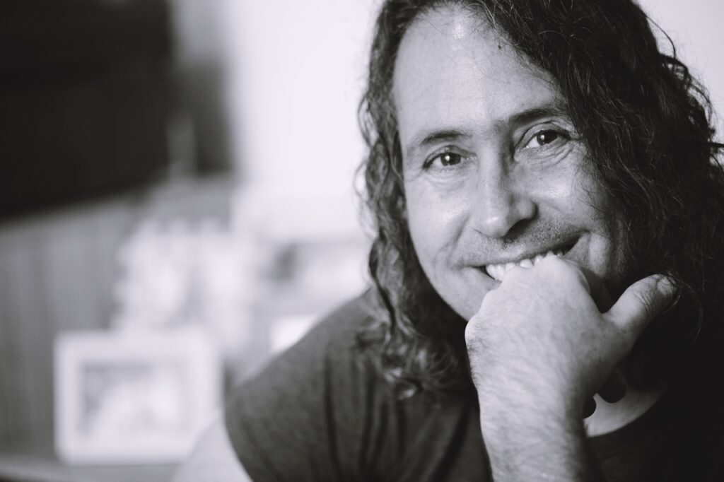 Retrato em preto e branco de um homem sorrindo, apoiando o queixo na mão, com cabelo comprido e ondulado.