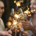 Close-up em primeiro plano de várias varetas de faíscas (sparklers) acesas, soltando brilhos dourados e intensos. Ao fundo, de forma desfocada, duas mulheres sorriem e celebram enquanto seguram garrafas de bebida. A iluminação é quente e festiva, sugerindo uma comemoração noturna ao ar livre.