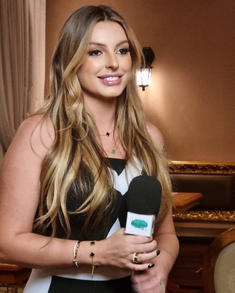 A empresária Muriel Prendin, reconhecida como uma das melhores maquiadoras do Brasil, sorri com confiança enquanto segura um pincel de sua marca própria próximo ao rosto. Ela está com um penteado sofisticado em coque alto e veste um traje elegante em preto e branco, simbolizando a maturidade e o sucesso de sua trajetória que começou em 2013 e culminou no lançamento de sua grife autoral em 2025.