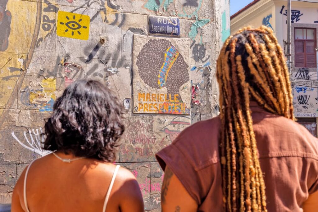 Larissa Amorim, nova Diretora de Programas e Estratégia do Instituto Marielle Franco, sorri sentada em uma escadaria pintada com a bandeira do Brasil. Ela segura a placa da "Rua Marielle Franco", simbolizando seu compromisso em fortalecer o legado da vereadora e atuar na defesa da democracia. A imagem une a nova liderança da organização ao propósito político e social de preservação da memória e fortalecimento de territórios defendido pelo Instituto.