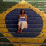 Uma mulher jovem, de pele negra e cabelos cacheados curtos, sorri sentada no centro de uma escadaria pintada com as cores e o design da bandeira do Brasil (verde, amarelo e azul). Ela usa um vestido bege claro e sandálias de tiras grossas na cor terracota. Em suas mãos, ela segura uma placa de rua azul e branca onde se lê "Rua Marielle Franco", contendo também informações biográficas sobre a vereadora e defensora dos direitos humanos. O ambiente é iluminado pela luz natural do sol, criando um contraste vibrante entre as cores da pintura e o cenário urbano.