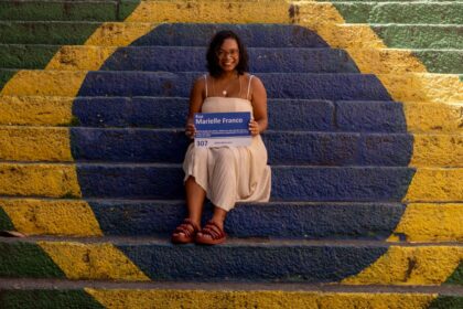 Uma mulher jovem, de pele negra e cabelos cacheados curtos, sorri sentada no centro de uma escadaria pintada com as cores e o design da bandeira do Brasil (verde, amarelo e azul). Ela usa um vestido bege claro e sandálias de tiras grossas na cor terracota. Em suas mãos, ela segura uma placa de rua azul e branca onde se lê "Rua Marielle Franco", contendo também informações biográficas sobre a vereadora e defensora dos direitos humanos. O ambiente é iluminado pela luz natural do sol, criando um contraste vibrante entre as cores da pintura e o cenário urbano.