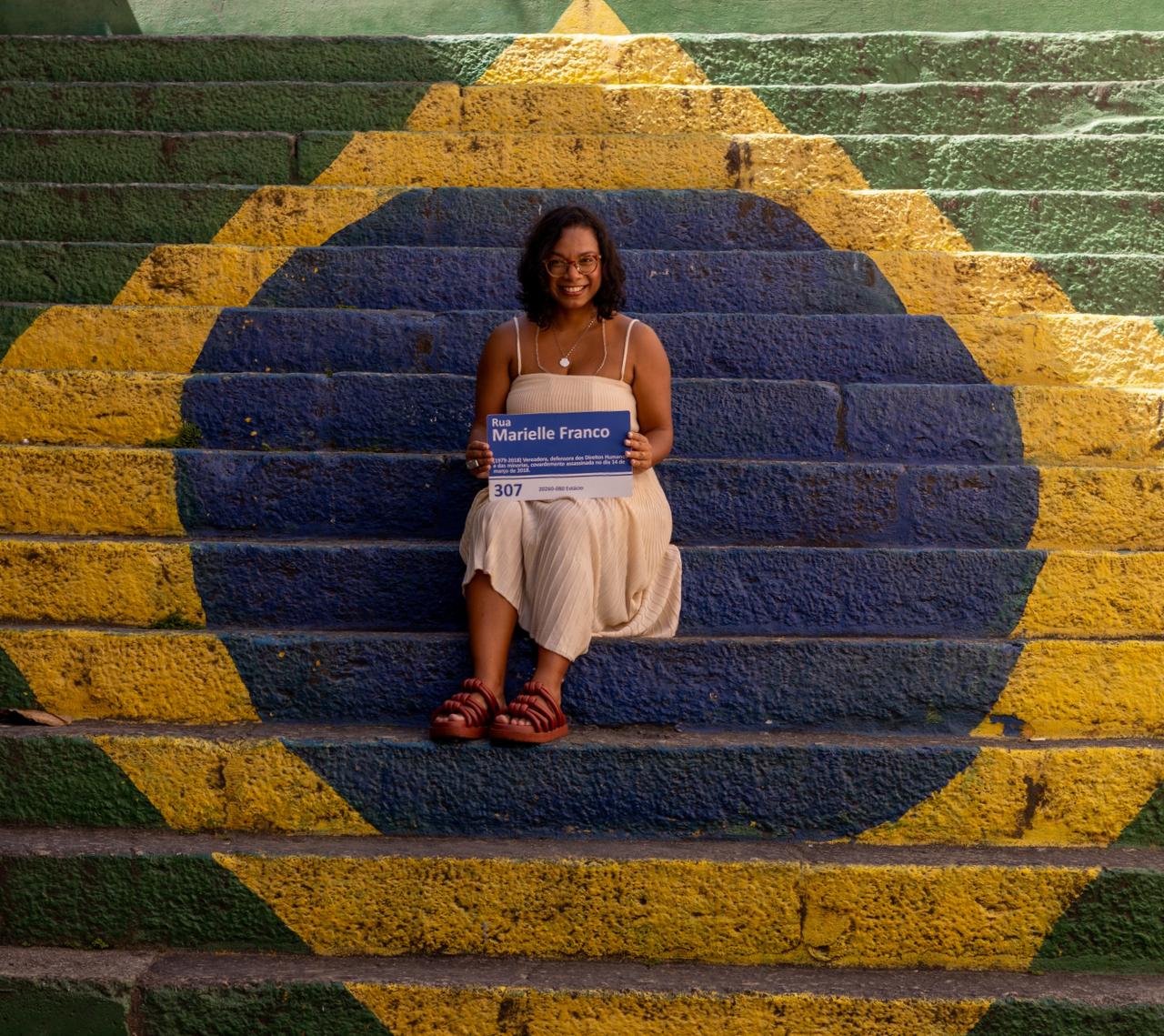 Uma mulher jovem, de pele negra e cabelos cacheados curtos, sorri sentada no centro de uma escadaria pintada com as cores e o design da bandeira do Brasil (verde, amarelo e azul). Ela usa um vestido bege claro e sandálias de tiras grossas na cor terracota. Em suas mãos, ela segura uma placa de rua azul e branca onde se lê "Rua Marielle Franco", contendo também informações biográficas sobre a vereadora e defensora dos direitos humanos. O ambiente é iluminado pela luz natural do sol, criando um contraste vibrante entre as cores da pintura e o cenário urbano.