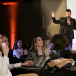 Homem de pé em um palco, segurando um microfone e levantando a mão enquanto apresenta uma palestra. À frente, várias pessoas sentadas acompanham atentamente, algumas usando notebooks e outras segurando copos de café, em um ambiente de evento ou conferência com iluminação quente e projeção ao fundo.