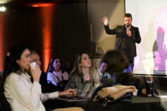 Homem de pé em um palco, segurando um microfone e levantando a mão enquanto apresenta uma palestra. À frente, várias pessoas sentadas acompanham atentamente, algumas usando notebooks e outras segurando copos de café, em um ambiente de evento ou conferência com iluminação quente e projeção ao fundo.