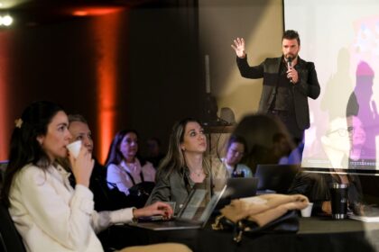 Homem de pé em um palco, segurando um microfone e levantando a mão enquanto apresenta uma palestra. À frente, várias pessoas sentadas acompanham atentamente, algumas usando notebooks e outras segurando copos de café, em um ambiente de evento ou conferência com iluminação quente e projeção ao fundo.