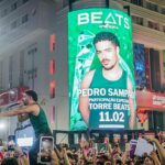 Cantor se apresenta em cima de um trio elétrico durante festa de Carnaval à noite, enquanto uma multidão registra o momento com celulares erguidos. Ao fundo, um grande telão verde exibe a imagem promocional de um artista e a frase “Beats apresenta” com data do evento, iluminando a fachada de um prédio e a avenida lotada.