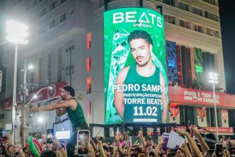 Cantor se apresenta em cima de um trio elétrico durante festa de Carnaval à noite, enquanto uma multidão registra o momento com celulares erguidos. Ao fundo, um grande telão verde exibe a imagem promocional de um artista e a frase “Beats apresenta” com data do evento, iluminando a fachada de um prédio e a avenida lotada.