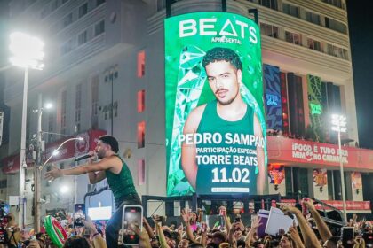 Cantor se apresenta em cima de um trio elétrico durante festa de Carnaval à noite, enquanto uma multidão registra o momento com celulares erguidos. Ao fundo, um grande telão verde exibe a imagem promocional de um artista e a frase “Beats apresenta” com data do evento, iluminando a fachada de um prédio e a avenida lotada.