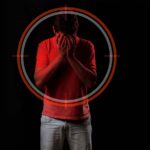 Foto de um homem jovem escondendo o rosto com as mãos, expressando angústia ou tristeza. Ele veste uma camiseta vermelha e calça clara, destacando-se contra um fundo preto. Sobre o seu tronco e cabeça, há a sobreposição gráfica de um alvo circular em tons de vermelho e cinza.