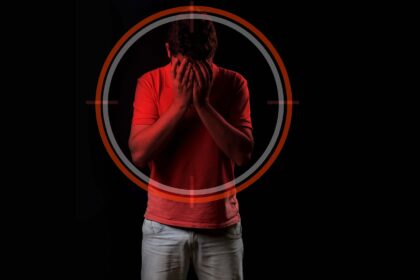 Foto de um homem jovem escondendo o rosto com as mãos, expressando angústia ou tristeza. Ele veste uma camiseta vermelha e calça clara, destacando-se contra um fundo preto. Sobre o seu tronco e cabeça, há a sobreposição gráfica de um alvo circular em tons de vermelho e cinza.